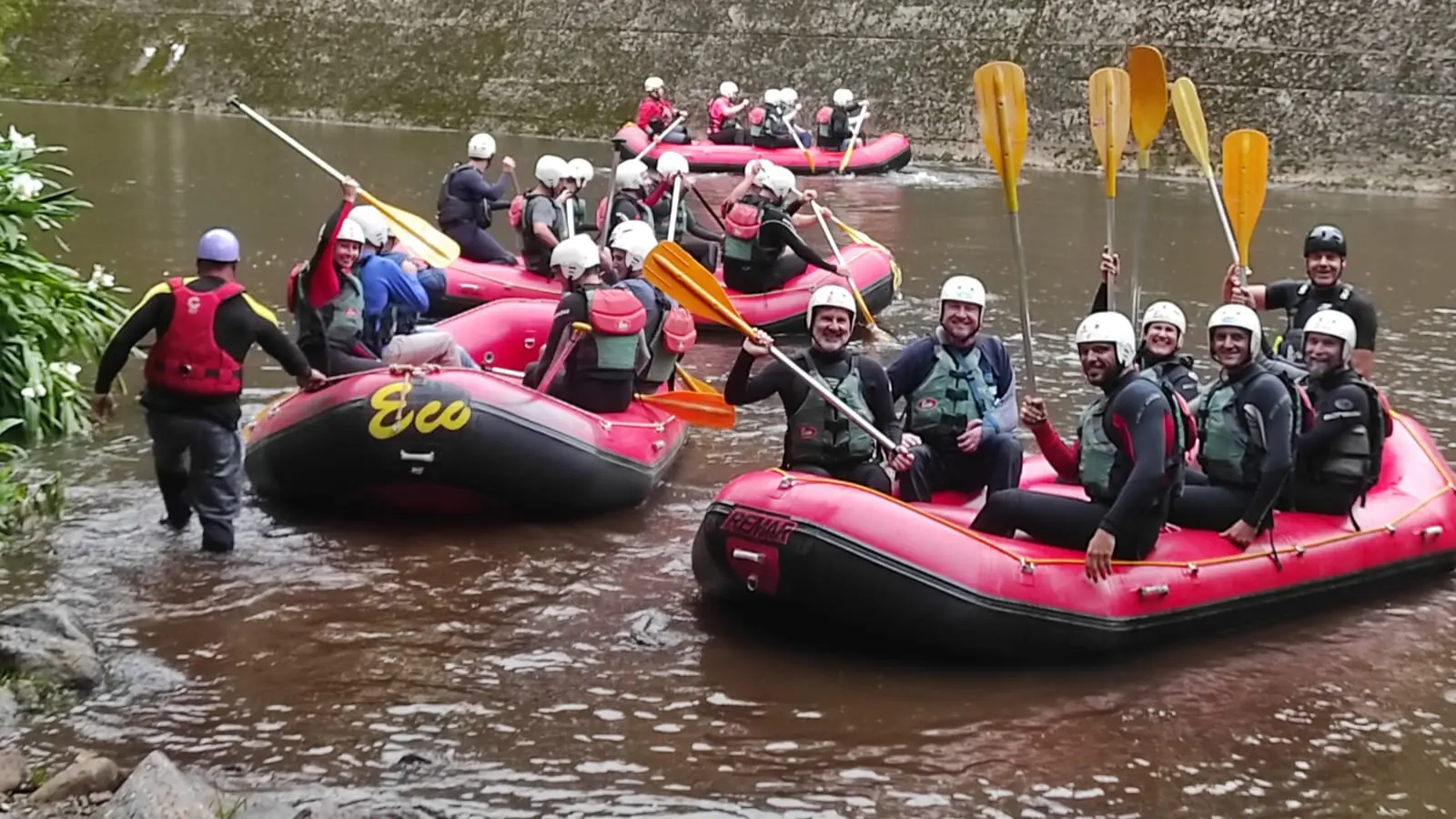 quatro botes de rafting na represa, repletos de pessoas segurando os remos, sorrindo e olhando para frente.